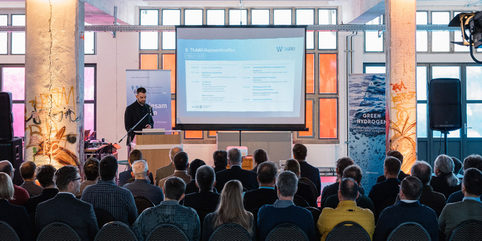 Präsentation auf dem ThAWI Netzwerktreffen im Kontor Erfurt 2026 Die Szene wurde von hinten fotografiert, es sitzen ca. 150 Leute im Raum vor einer Leinwand auf der eine Präsentation von Robert Kraft auf dem ThAWI Netzwerktreffen im Kontor Erfurt 2026 läuft. Robert Kraft der ThEGA steht auf der Bühne.