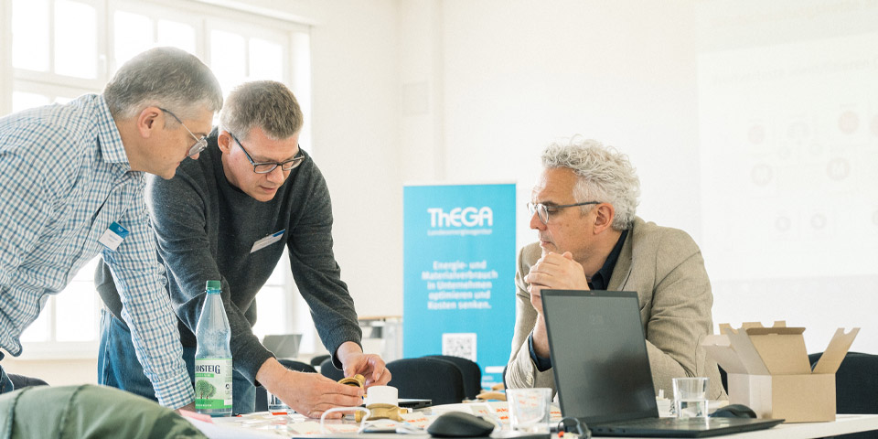 Workshop Gespräch von drei Personen In einem hellen Raum reden drei Personen über ein fachlichen Sachverhalt, zwei Personen stehen und erklären, die dritte Person sitzt und hört zu.